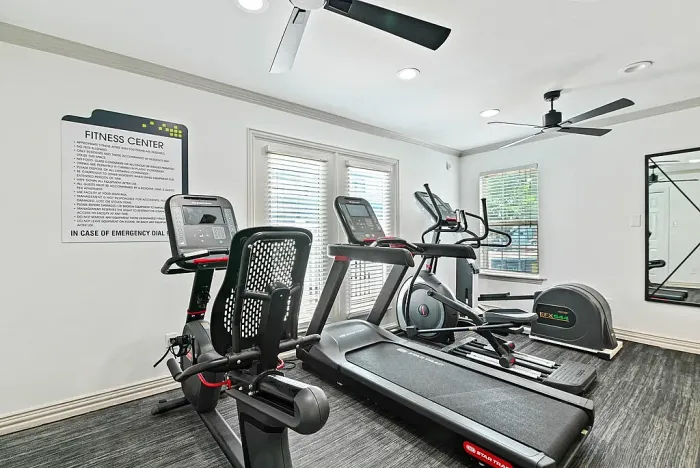 Fitness center with cardio equipment including a treadmill, elliptical machines, and a recumbent bike. The space features large windows with natural light, ceiling fans, a wall mirror, and posted fitness guidelines, creating a bright and functional workout area.