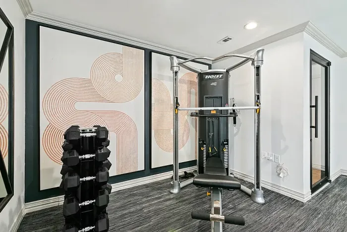 Fitness center with a cable weight machine, adjustable bench, and a rack of dumbbells. The space features modern wall art, mirrors, neutral walls, and dark flooring, creating a clean and functional workout environment.