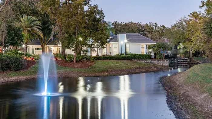 Scenic waterfront pond with illuminated fountain, walking path, and landscaped grounds at dusk.