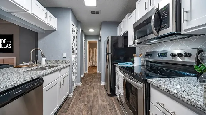 Galley-style kitchen with granite countertops, white cabinets, stainless steel appliances, subway tile backsplash, and wood-style flooring leading to a hallway.