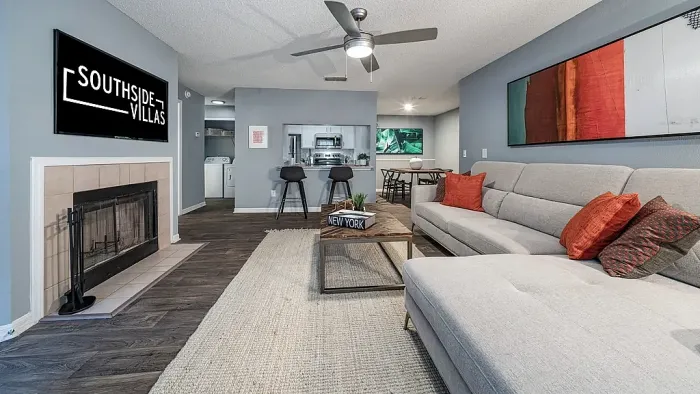 Spacious living room with wood-style flooring, a large sectional sofa, ceiling fan, and fireplace, opening to a kitchen with breakfast bar and adjacent dining area.