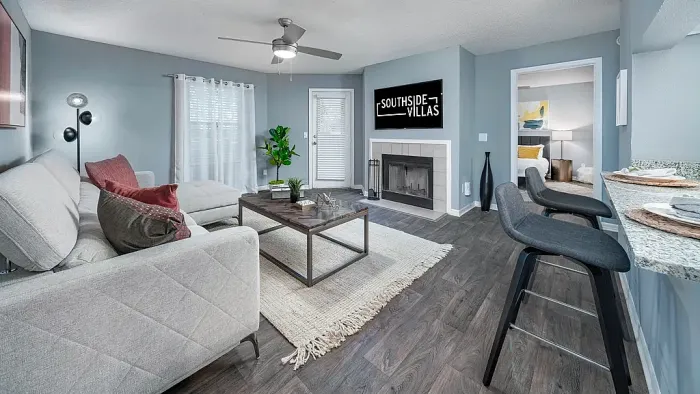 Bright living room with wood-style flooring, ceiling fan, and fireplace, featuring a sofa seating area and breakfast bar seating, with access to a bedroom and outdoor patio.