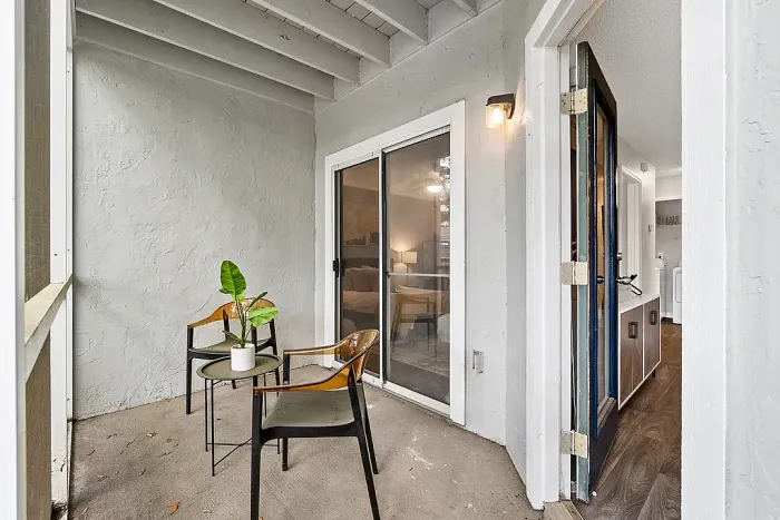 Covered patio with two chairs and a small table, featuring sliding glass door access to the bedroom and entry into the apartment.
