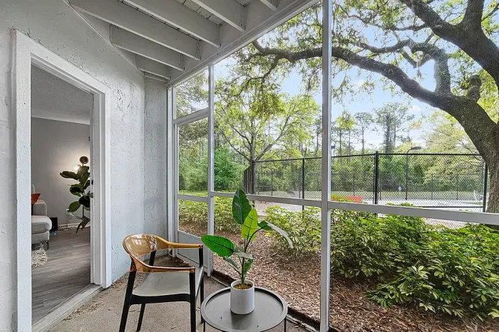 Screened-in patio with seating and a small table, overlooking landscaped greenery and a nearby tennis court.