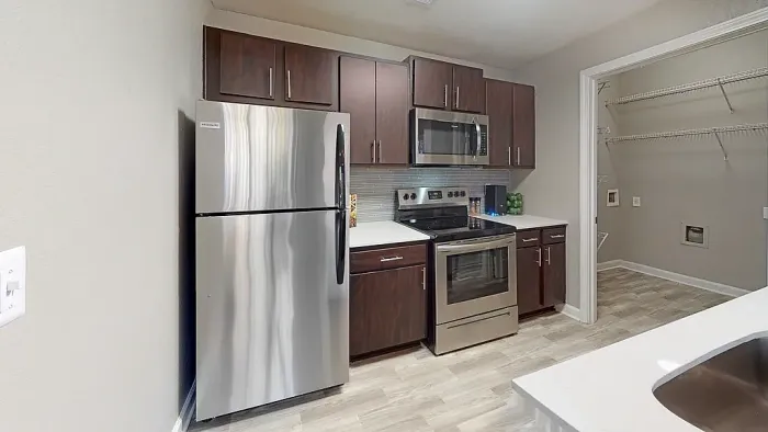 Modern apartment kitchen featuring dark wood cabinets, stainless steel appliances, white quartz-style countertops, gray subway tile backsplash, wood-style flooring, and an adjacent laundry/storage area with built-in shelving.