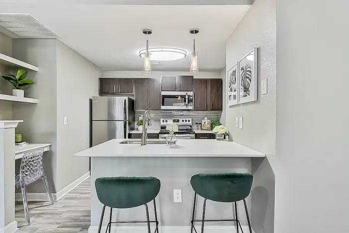 Modern apartment kitchen with dark wood cabinetry, stainless steel appliances, white quartz-style island with seating for two, pendant lighting, gray tile backsplash, and open shelving in a bright open-concept layout.