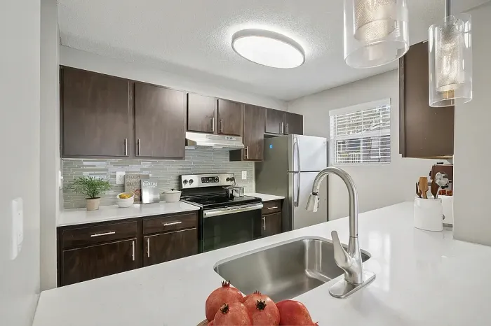 Modern apartment kitchen with dark wood cabinetry, stainless steel appliances, white quartz-style countertops, gray glass tile backsplash, pendant lighting, and a large island sink with contemporary finishes.