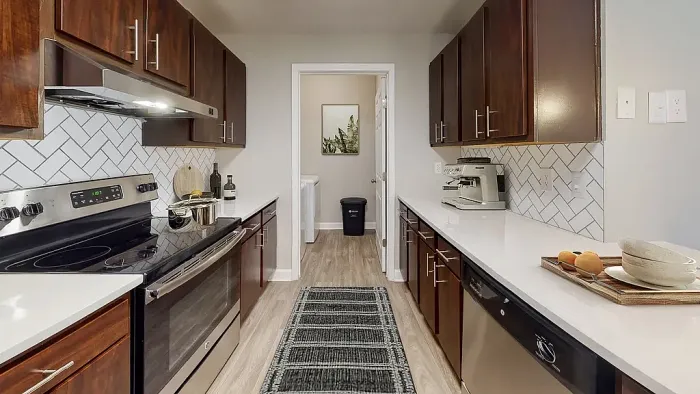 Modern galley-style apartment kitchen with dark wood cabinets, stainless steel appliances, white quartz-style countertops, white herringbone tile backsplash, wood-style flooring, and a spacious open layout leading to a laundry area.