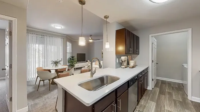Modern open-concept apartment kitchen with dark wood cabinetry, stainless steel appliances, white quartz-style countertops, pendant lighting, and a large breakfast bar overlooking the dining and living areas.