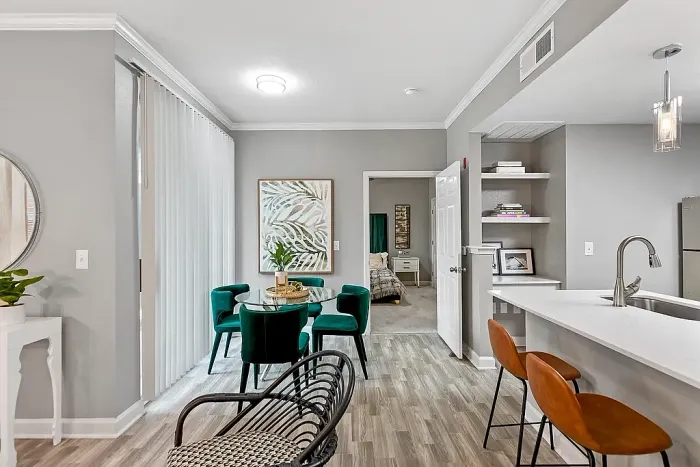 Modern open-concept apartment dining and kitchen area with wood-style flooring, a breakfast bar with seating, built-in shelving, pendant lighting, and a dining nook with emerald green velvet chairs and contemporary decor.