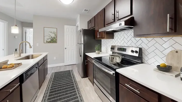 Modern galley-style apartment kitchen with dark wood cabinetry, stainless steel appliances, white quartz-style countertops, white herringbone tile backsplash, pendant lighting, and wood-style flooring.