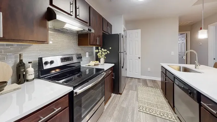 Modern galley-style apartment kitchen with dark wood cabinetry, stainless steel appliances, white quartz-style countertops, gray glass tile backsplash, pendant lighting, and wood-style flooring with a spacious island sink.