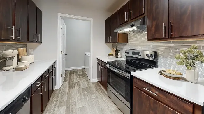 Modern galley-style apartment kitchen with dark wood cabinetry, stainless steel appliances, white quartz-style countertops, gray tile backsplash, and wood-style flooring with contemporary finishes throughout.