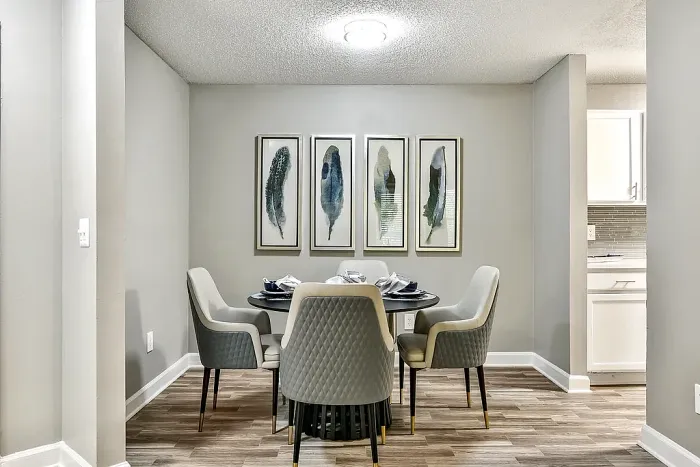 Modern apartment dining area with wood-style flooring, a round dining table with upholstered seating for four, neutral gray walls, and contemporary abstract wall art.