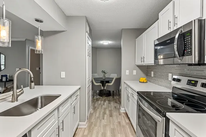 Modern galley-style apartment kitchen with white shaker cabinetry, stainless steel appliances, gray tile backsplash, quartz-style countertops, pendant lighting, and wood-style flooring leading into a dining area.