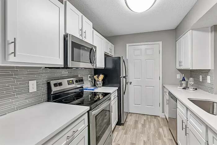Modern galley-style apartment kitchen with white shaker cabinets, stainless steel appliances, gray glass tile backsplash, quartz-style countertops, and wood-style flooring with contemporary finishes.