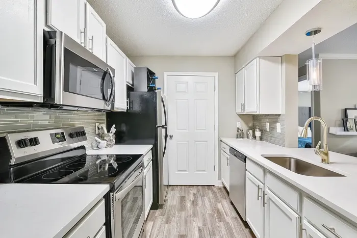 Modern galley-style apartment kitchen with white shaker cabinetry, stainless steel appliances, quartz-style countertops, gray glass tile backsplash, pendant lighting, and wood-style flooring.