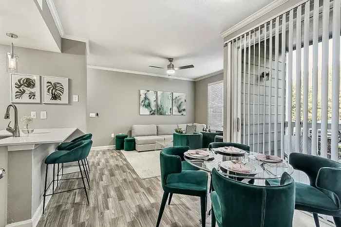 Modern open-concept apartment living and dining area with emerald green velvet seating, wood-style flooring, a breakfast bar with pendant lighting, large sliding glass doors, and contemporary decor throughout.
