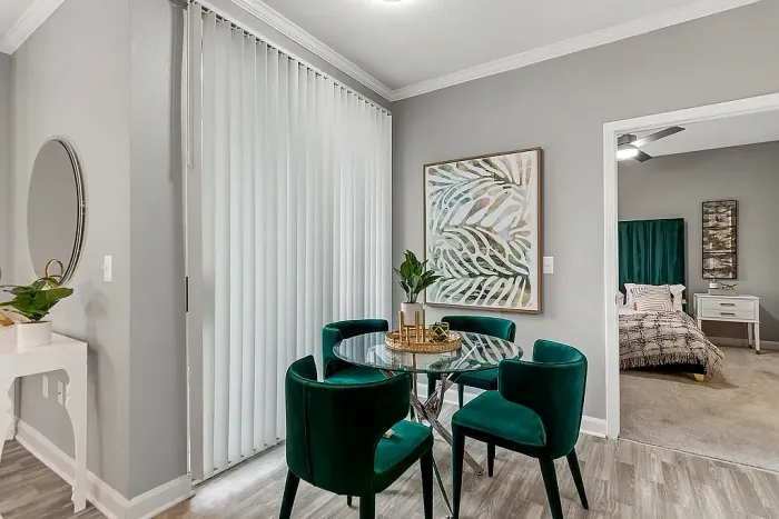 Modern apartment dining nook with a glass-top dining table, emerald green velvet chairs, contemporary wall art, wood-style flooring, and an open view into the bedroom area.