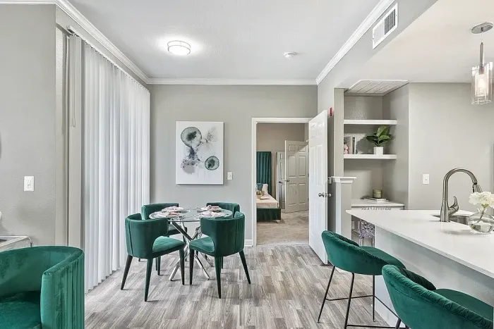 Modern open-concept apartment dining and kitchen area with emerald green velvet seating, a breakfast bar with quartz-style countertops, built-in shelving, wood-style flooring, and contemporary decor throughout.