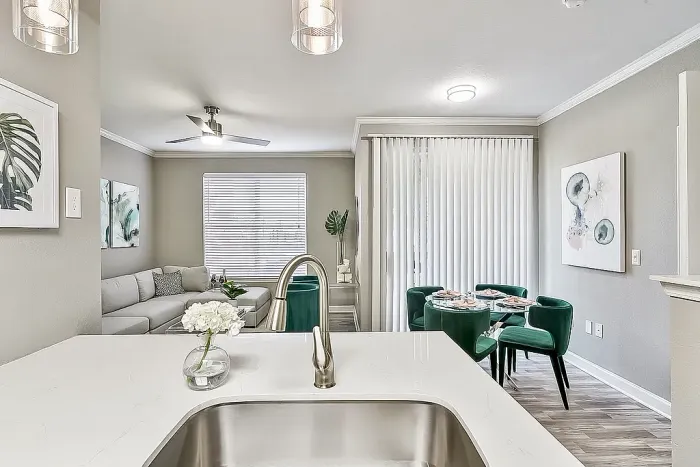 Modern open-concept apartment kitchen and living area with quartz-style countertops, pendant lighting, emerald green velvet dining chairs, wood-style flooring, and contemporary furnishings throughout.