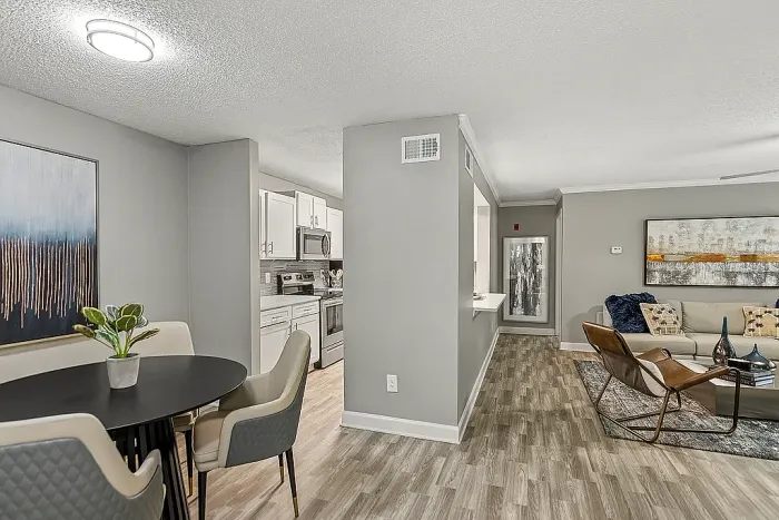 Modern open-concept apartment dining and living area with wood-style flooring, neutral gray tones, contemporary furnishings, abstract wall art, and a view into the updated kitchen with white cabinetry and stainless steel appliances.