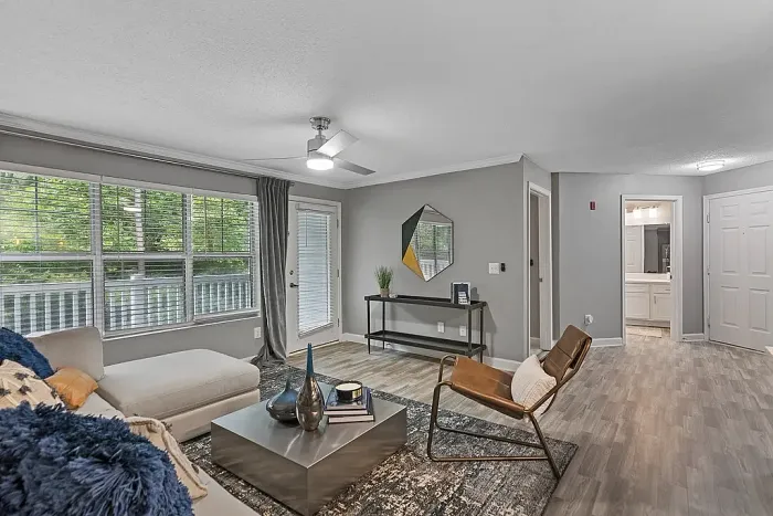 Modern apartment living room with wood-style flooring, large windows for natural light, contemporary furnishings, neutral gray walls, and access to a private patio or balcony.