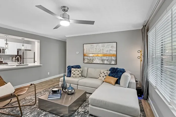 Modern apartment living room with wood-style flooring, a sectional sofa, contemporary artwork, large windows with blinds, and an open-concept layout connected to the kitchen area.
