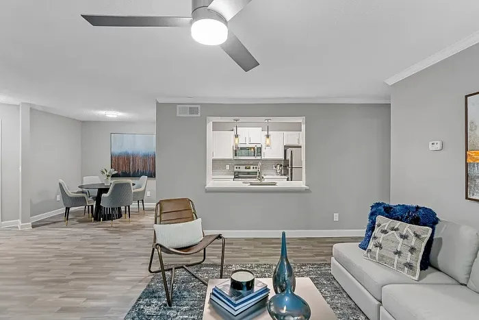 Spacious open-concept apartment living and dining area with wood-style flooring, contemporary furnishings, neutral gray tones, and a pass-through kitchen with white cabinetry and pendant lighting.