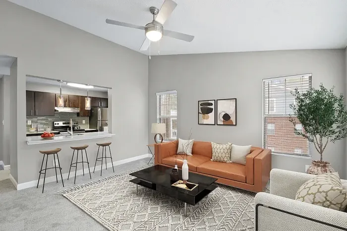 Modern apartment living room with vaulted ceilings, contemporary furnishings, neutral gray walls, large windows for natural light, and a breakfast bar opening into the kitchen with pendant lighting and seating.