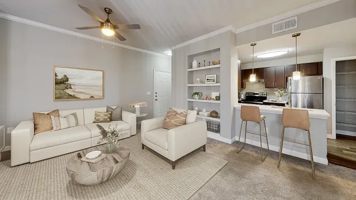 Modern open-concept apartment living room and kitchen with neutral furnishings, plush carpeting, built-in shelving, a breakfast bar with seating, stainless steel appliances, and contemporary finishes throughout.