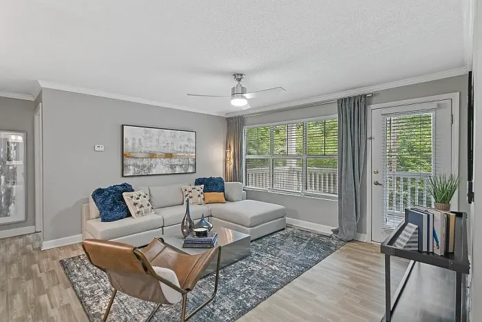 Modern apartment living room with wood-style flooring, large windows for natural light, contemporary furnishings, neutral gray tones, and access to a private balcony or patio.