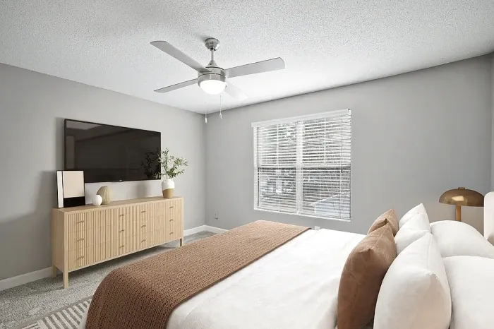 Modern apartment bedroom with plush carpeting, neutral gray walls, large windows for natural light, a ceiling fan, and contemporary furnishings with warm neutral accents.