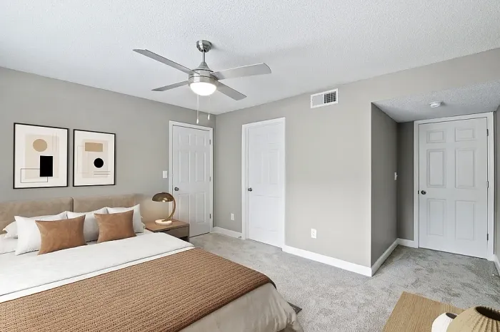 Modern apartment bedroom with plush carpeting, neutral gray walls, a ceiling fan, contemporary furnishings, and warm neutral decor accents with ample natural light.