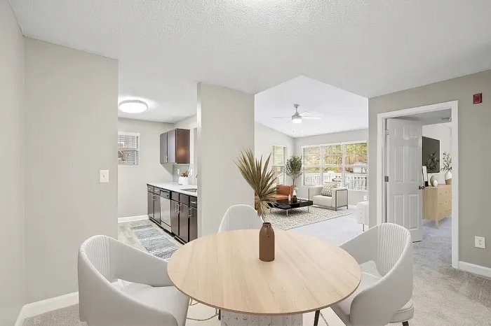 Modern open-concept apartment dining and living area with neutral tones, contemporary furnishings, a round wood dining table with upholstered seating, and views into the kitchen and bedroom spaces.