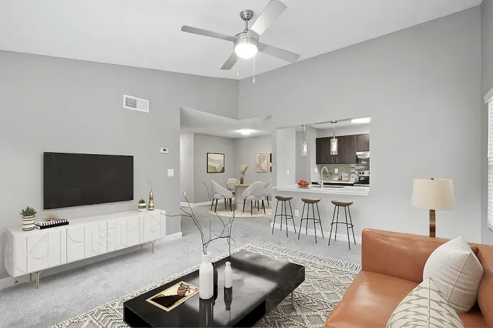 Modern open-concept apartment living room with vaulted ceilings, plush carpeting, contemporary furnishings, a breakfast bar with seating, and a pass-through kitchen with pendant lighting and stainless steel appliances.