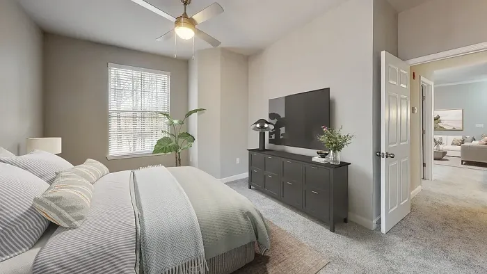 Modern apartment bedroom with plush carpeting, neutral tones, a ceiling fan, large window for natural light, and contemporary furnishings with an open view into the adjoining living area.