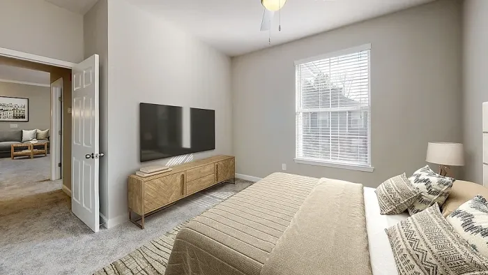 Modern apartment bedroom with plush carpeting, neutral tones, large window for natural light, contemporary furnishings, and an open doorway leading to the adjoining living area.