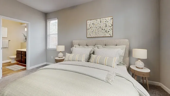 Cozy primary bedroom with plush carpeting, upholstered bed, soft neutral tones, matching bedside tables with lamps, and direct access to an en-suite bathroom.