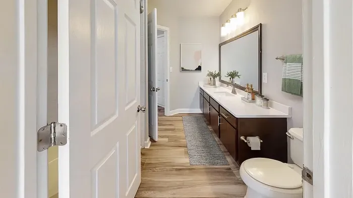 Spacious bathroom featuring a long dual-sink vanity, oversized framed mirror, modern lighting, wood-style flooring, and elegant dark wood cabinetry with ample storage.
