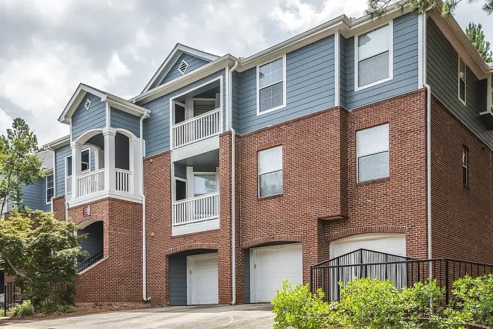 Three-story apartment community exterior featuring classic red brick architecture, blue siding accents, private balconies, attached garages, and beautifully maintained landscaping.