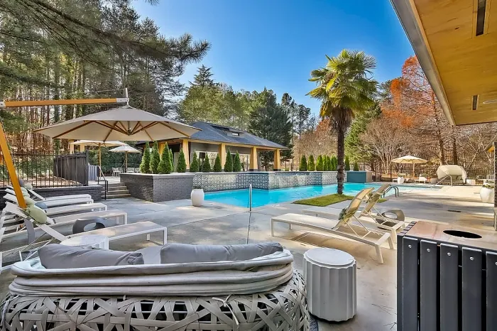 Luxurious resort-style pool area with expansive lounge seating, shaded cabanas, tropical landscaping, and a modern clubhouse surrounded by peaceful wooded views.