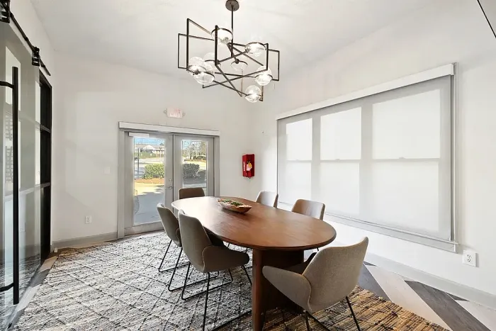 Modern private conference room with a long oval wood table, six upholstered chairs, a geometric black chandelier with glass globe lights, floor-to-ceiling windows with roller shades, and glass double doors leading outside.