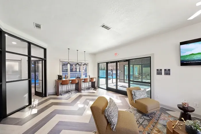 Bright modern resident lounge with geometric wood-look flooring, cozy lounge seating, a built-in work bar with pendant lighting and stools, floor-to-ceiling glass doors leading to the pool area, and contemporary black-framed interior windows and doors.