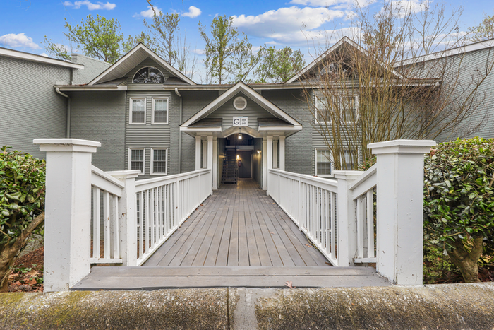 Charming apartment building entrance with a welcoming wooden walkway and lush greenery, surrounded by elegant gray brick architecture.