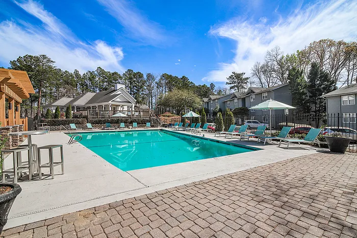 Luxurious apartment pool area featuring a sparkling swimming pool, stylish sun loungers, and scenic landscaping under a clear blue sky.