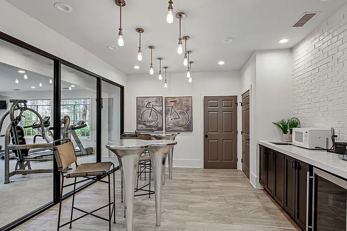 Modern communal kitchen with chic metal bar stools, contemporary lighting, and sleek cabinetry, adjacent to a well-equipped glass-walled fitness center.