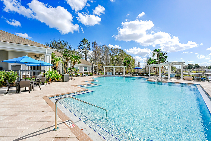 Inviting resort-style swimming pool with lush landscaping, poolside seating, and vibrant blue umbrellas under a sunny sky.
