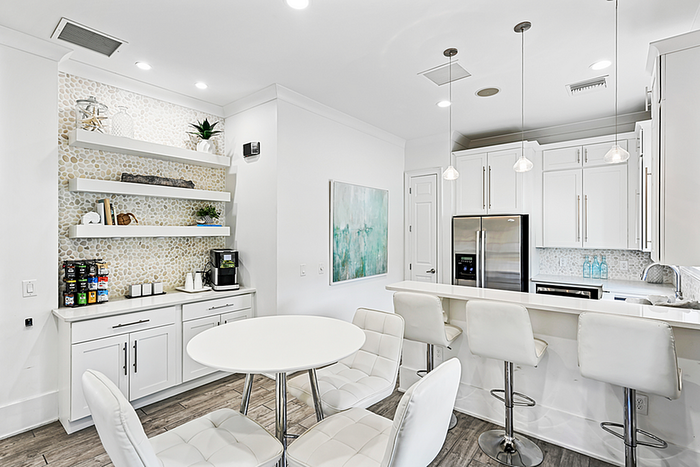 Elegant modern clubhouse kitchen with sleek white cabinets, pendant lights, and a chic breakfast bar. Open shelving adds a stylish touch in a bright, inviting space.