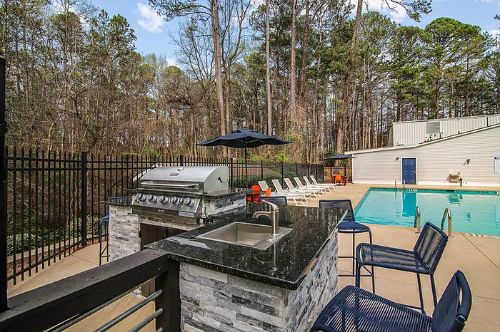 Inviting outdoor pool area featuring a modern grill station and comfortable lounge chairs, surrounded by lush woods.